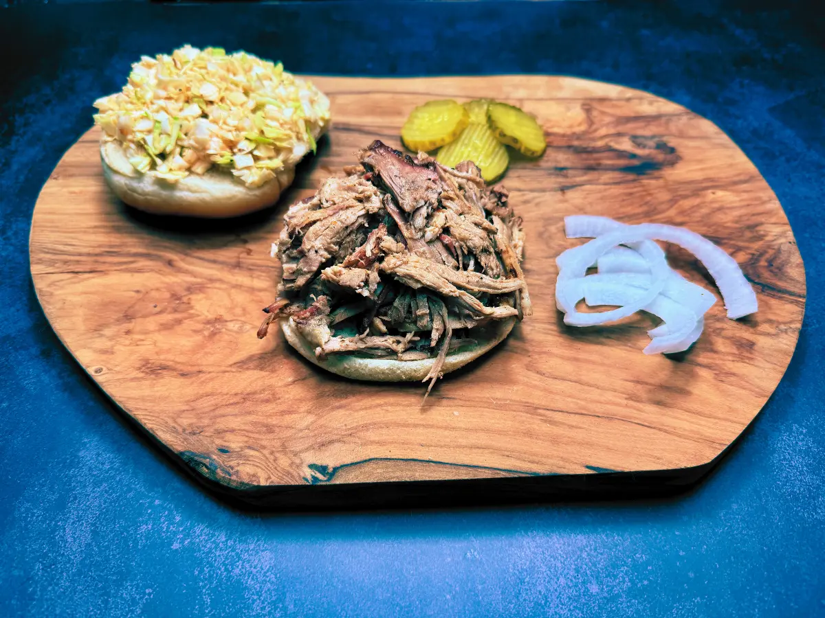 nicely displayed pulled pork sandwich on wooden cutting board with pickles and onions