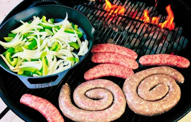 sausage and bratwurst cooking next to cast iron pan with peppers and onions on charcoal grill